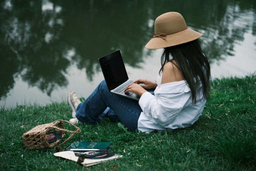 Woman in a sun hat sitting by the water, working on a laptop with a notebook and books beside her
